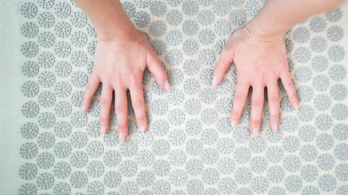 Nahaufnahme von zwei Händen, die auf einer Akupressurmatte liegen. Die Matte ist mit runden, stacheligen Massagepunkten ausgestattet, die zur Stimulation der Handflächen und Förderung der Durchblutung dienen. Die Person trägt ein schlichtes Armband und die Szene zeigt einen Moment der Entspannung oder Akupressurbehandlung.